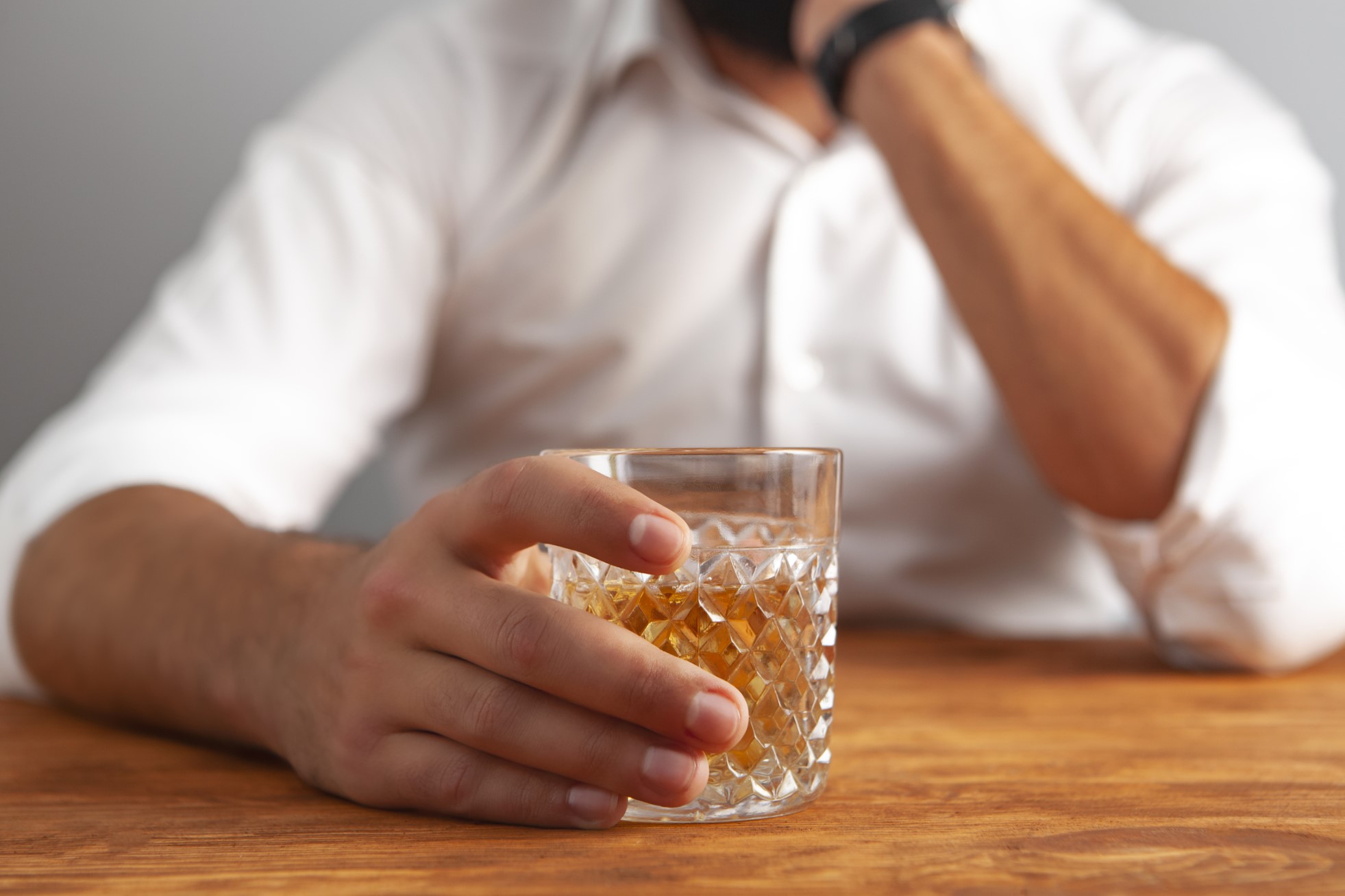 The image depicts a man, largely out of focus in the background, seated at a wooden table holding a glass of whiskey, evoking contemplation.