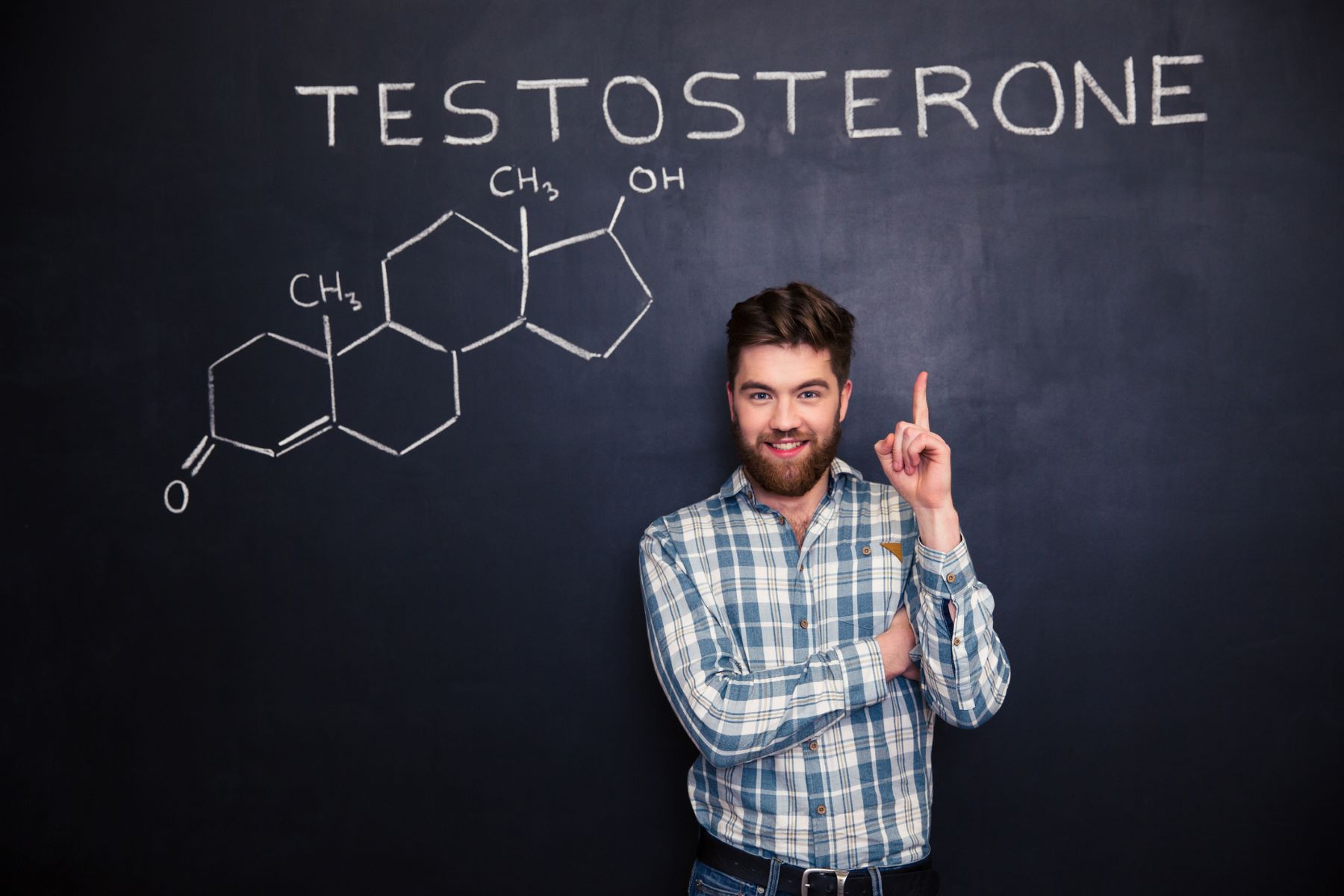A young man standing in front of a blackboard, pointing at the word “testosterone” written on it above his head.