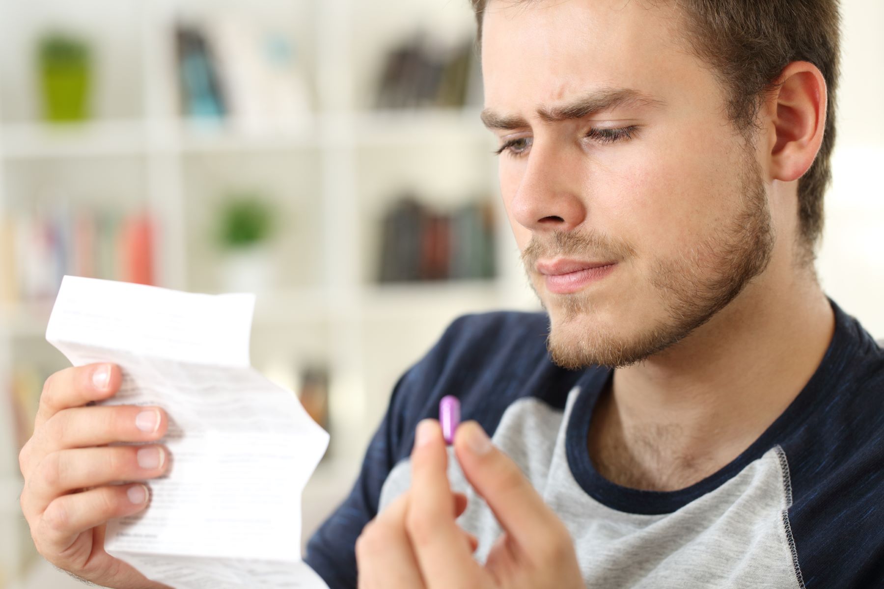 Young man holding a medication capsule and frowning while reading the package insert